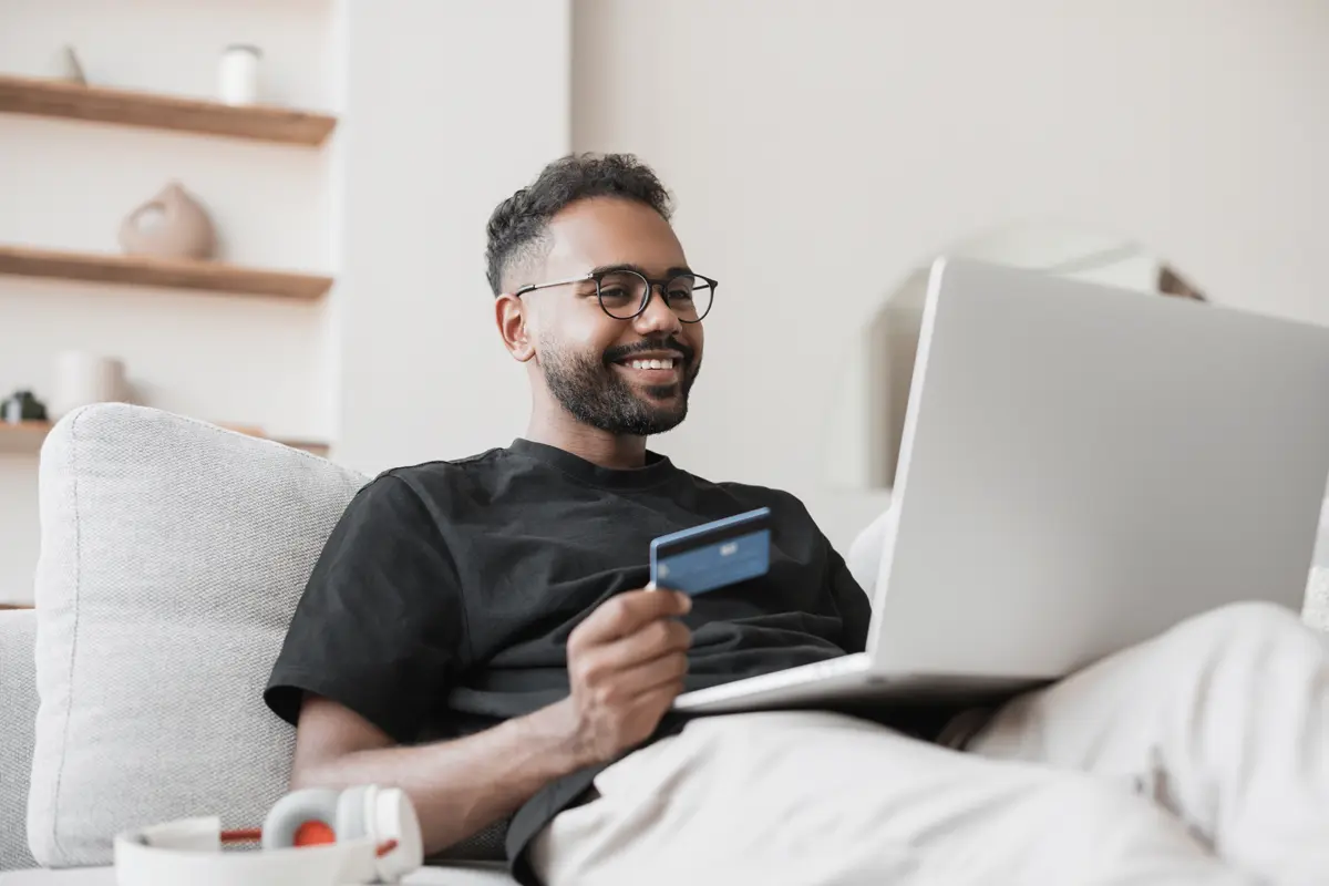man on a white sofa holding a laptop and a credit card
